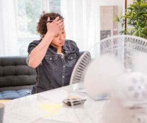 Lady sitting at her desk with hand on forehead, feeling hot and flustered. a fan is in the foreground placed on her desk.