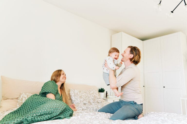 Young happy couple mother and father playing with adorable toddler 1 year old girl, family relaxing in bed