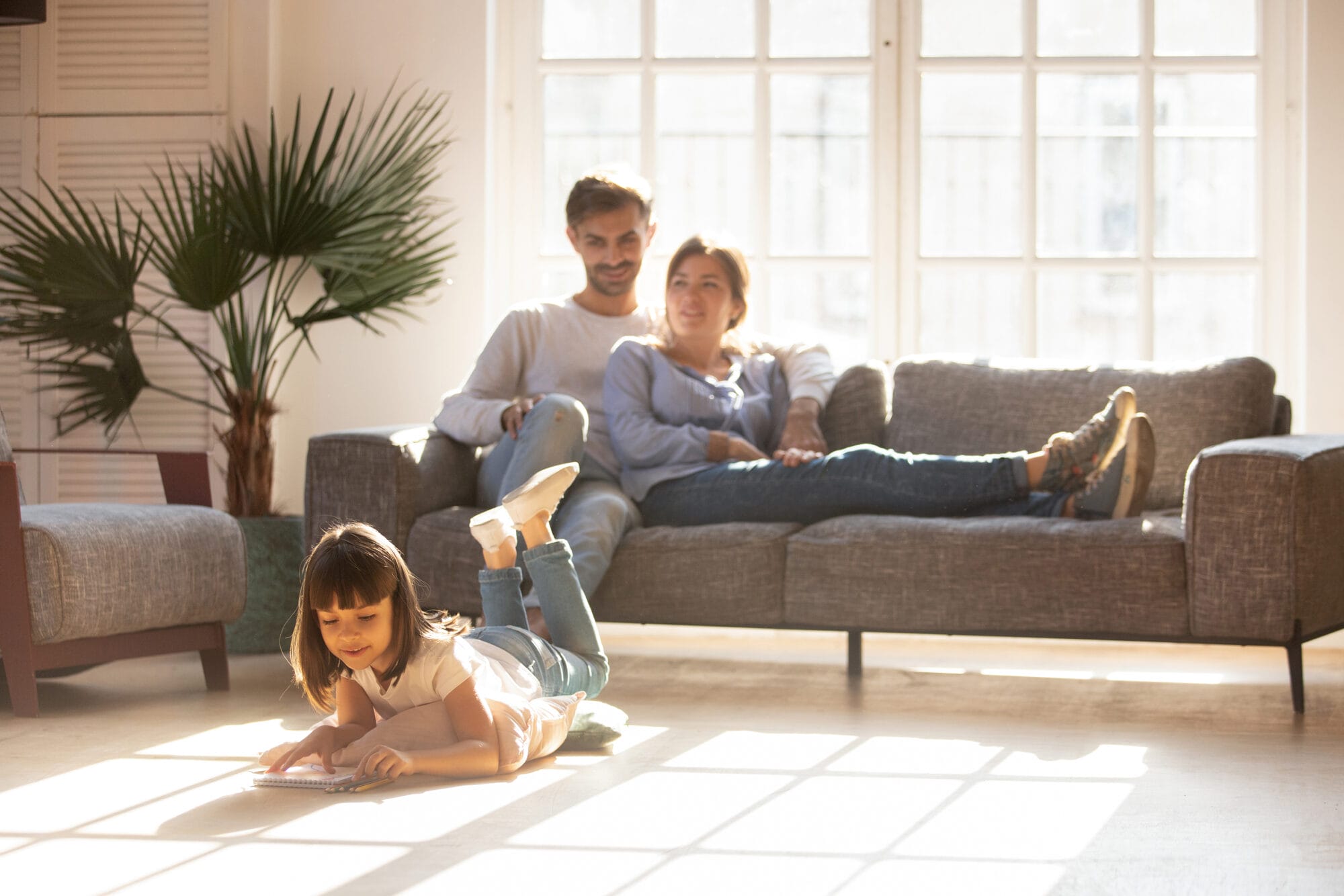 Family of three at home. Parents sitting on sofa with little girl playing on the floor.